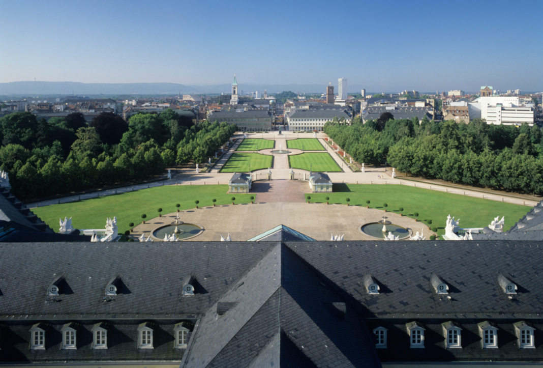 Über dem Dach des Karlsruher Schlosses fällt der Blick auf die Innenstadt von Karlsruhe. Im Vordergrund sind Park- und Waldanlagen zu erkennen sowie Teile des Schlossdaches.