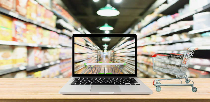 Dgitalisierung im Handel, dargestellt durch aufgeklappten Laptop zwischen Supermarktregalen. Laptopmonitor und Realität zeigen den Flur zwischen den Regalen und zeigen einen realen Einkaufwagen.