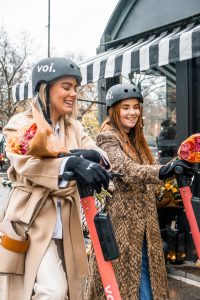 Hochkant-Foto mit zwei Frauen (blon und brünett) mit schwarzem Helm mit voi.-Logo und zwei E-Scootern (im Anschnitt), den die Frauen gerade schieben. Beide Frauen tragen einen langen Wintermantel und halten einen Strauß in Papier eingewickelter Blumen in der Hand. Die Frauen tragen zudem schwarze Handschuhe mit voi.-Logoaufdruck.